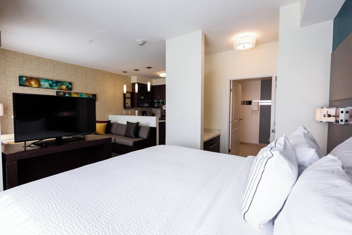 A hotel room with a large bed, a flat-screen TV, a sofa, and a kitchen area in the background.