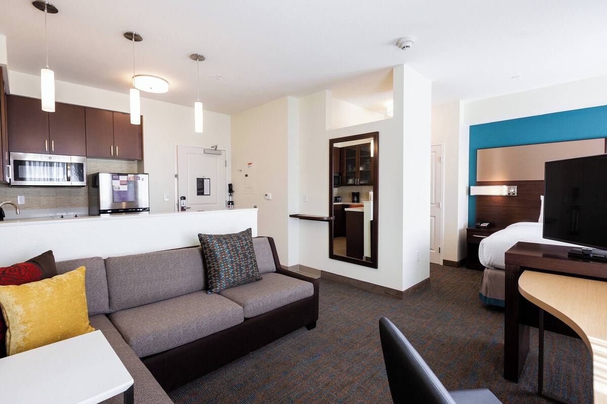 A modern hotel room with a kitchenette, a sofa with cushions, a dining table, and a TV.
