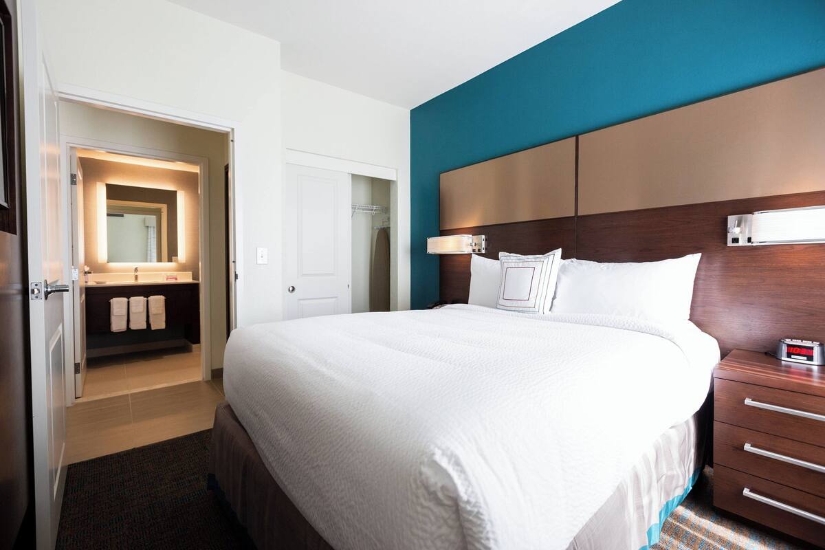 A hotel room with a large bed, a wooden headboard, a nightstand with a clock, and a bathroom visible through an open door.