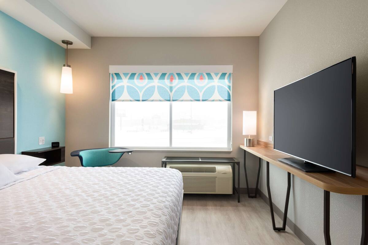 Ein Hotelzimmer mit einem Bett, einem Schreibtisch mit Flachbildfernseher, einem Sessel, einem Fenster mit gemusterten Vorhängen und einem Heizkörper.