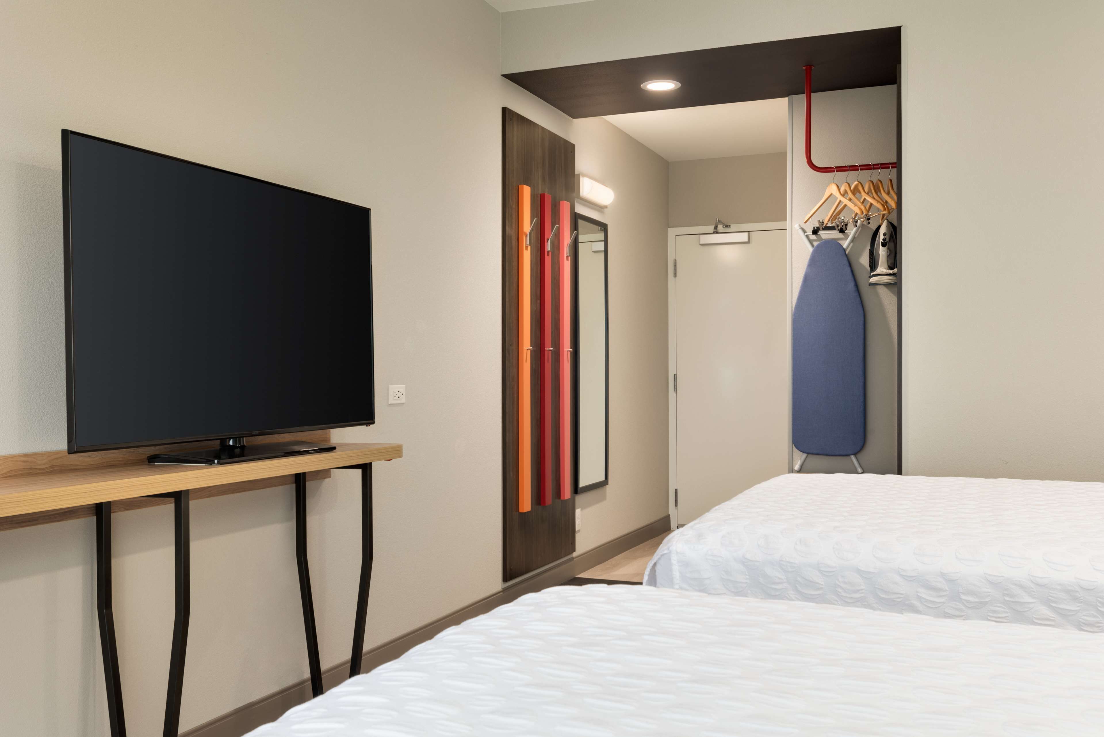 Une chambre d’hôtel équipée d’un téléviseur à écran plat posé sur un meuble en bois, d’un miroir à rayures verticales, d’une porte et d’une planche à repasser avec des cintres.