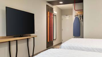 Une chambre d’hôtel équipée d’un téléviseur à écran plat posé sur un meuble en bois, d’un miroir à rayures verticales, d’une porte et d’une planche à repasser avec des cintres.