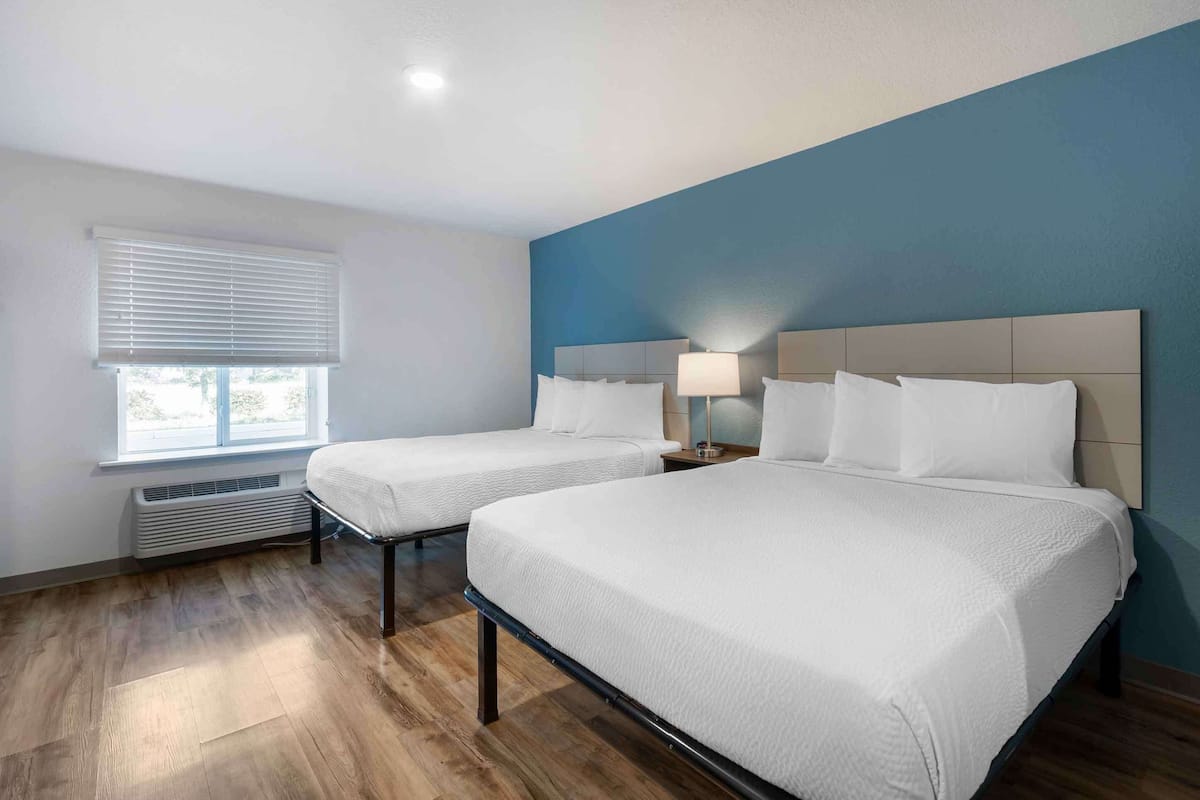Una habitación de hotel con dos camas, una ventana con persianas, una mesita de noche con lámpara y un calentador de pared.