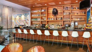Zona de bar con una barra larga, taburetes blancos y una variedad de botellas y vasos en estantes.