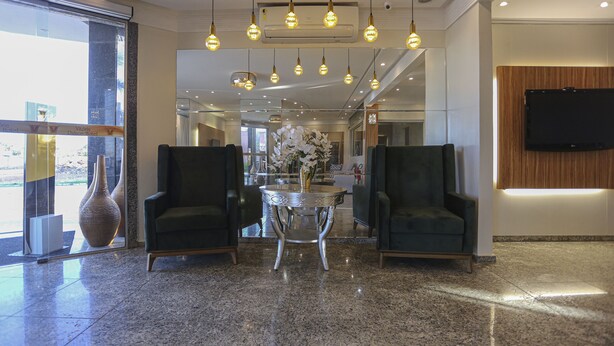 A lobby with two dark armchairs, a round table with a vase, and a mounted TV. - Vitoria Palace Hotel (Mossoro)