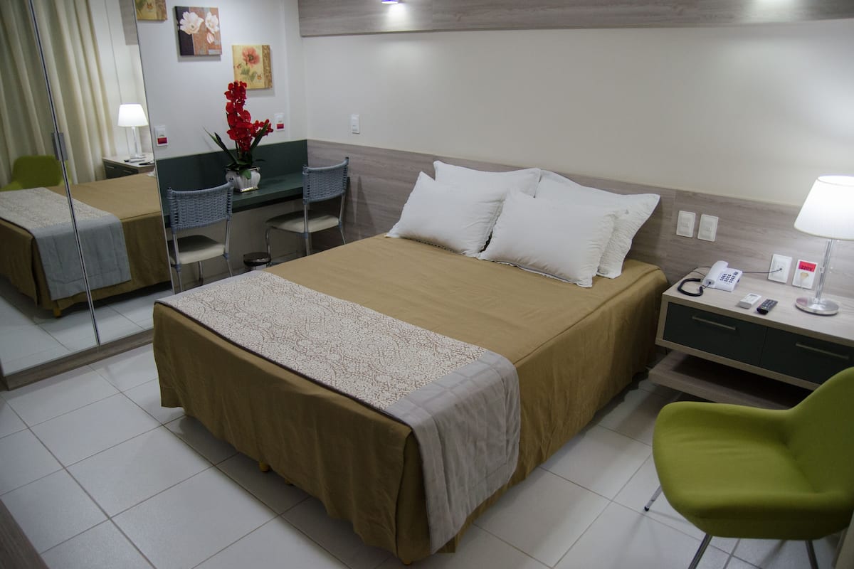 A hotel room with a large bed, a desk, a chair, and a small table with a lamp.