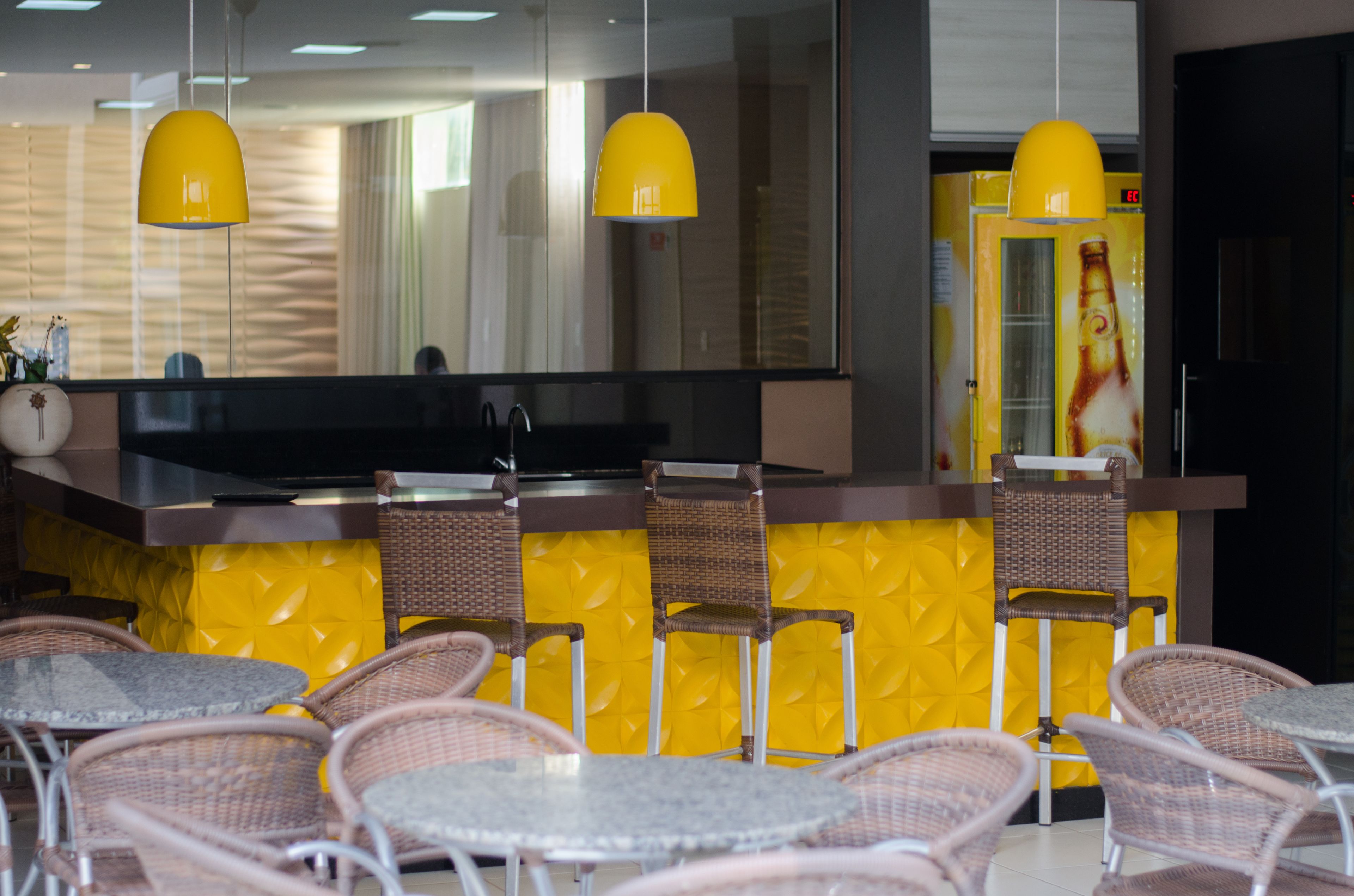 Un restaurant aux murs jaunes, avec des chaises en osier et un coin bar doté d’un grand écran.