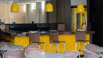 A restaurant with yellow walls, wicker chairs, and a bar area with a large screen.