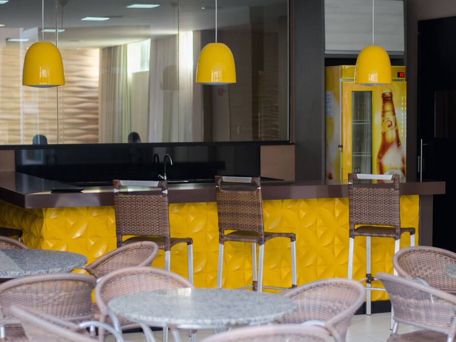 A restaurant with yellow walls, wicker chairs, and a bar area with a large screen.