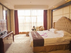 Ein Hotelzimmer mit einem großen Bett, einem Fernseher, einem Schreibtisch und einem Fenster mit Vorhängen.