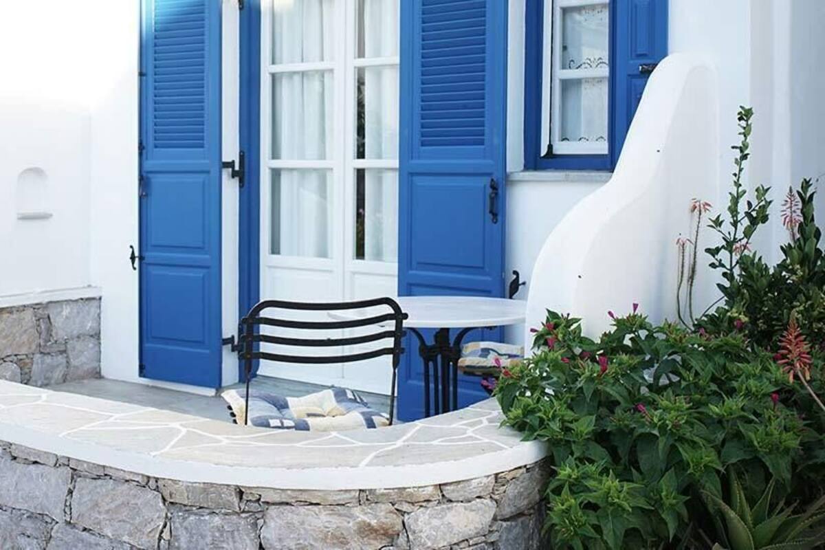 Un muro de piedra, una mesa blanca con una silla negra y una puerta azul con contraventanas.