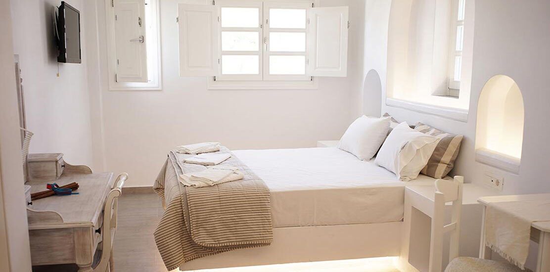 Ein Schlafzimmer mit einem Bett, Nachttischen, einem Schreibtisch, einem Sessel, einem Fenster mit Fensterläden und einem an der Wand montierten Fernseher.
