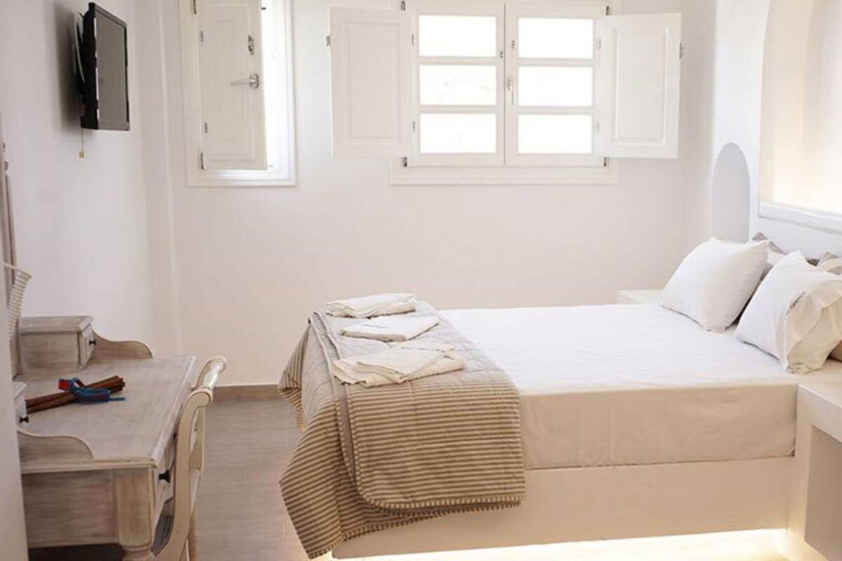 Un dormitorio con cama, mesitas de noche, escritorio, silla, ventana con contraventanas y televisor en la pared.