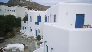 A row of white buildings with blue doors and windows, a stone courtyard with seating, and a mountain in the background. - Paraporti Hotel (Folegandros)