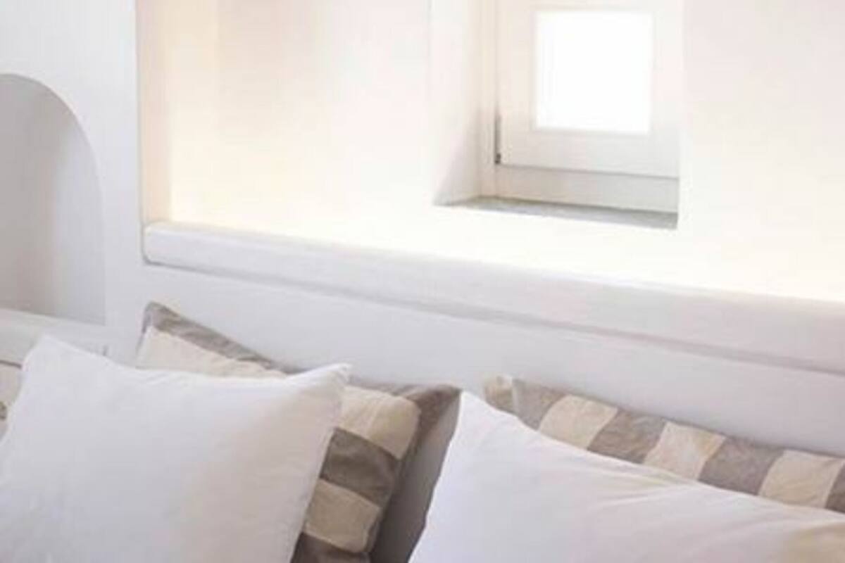 Un dormitorio con ventana, cama con almohadas y pared blanca.