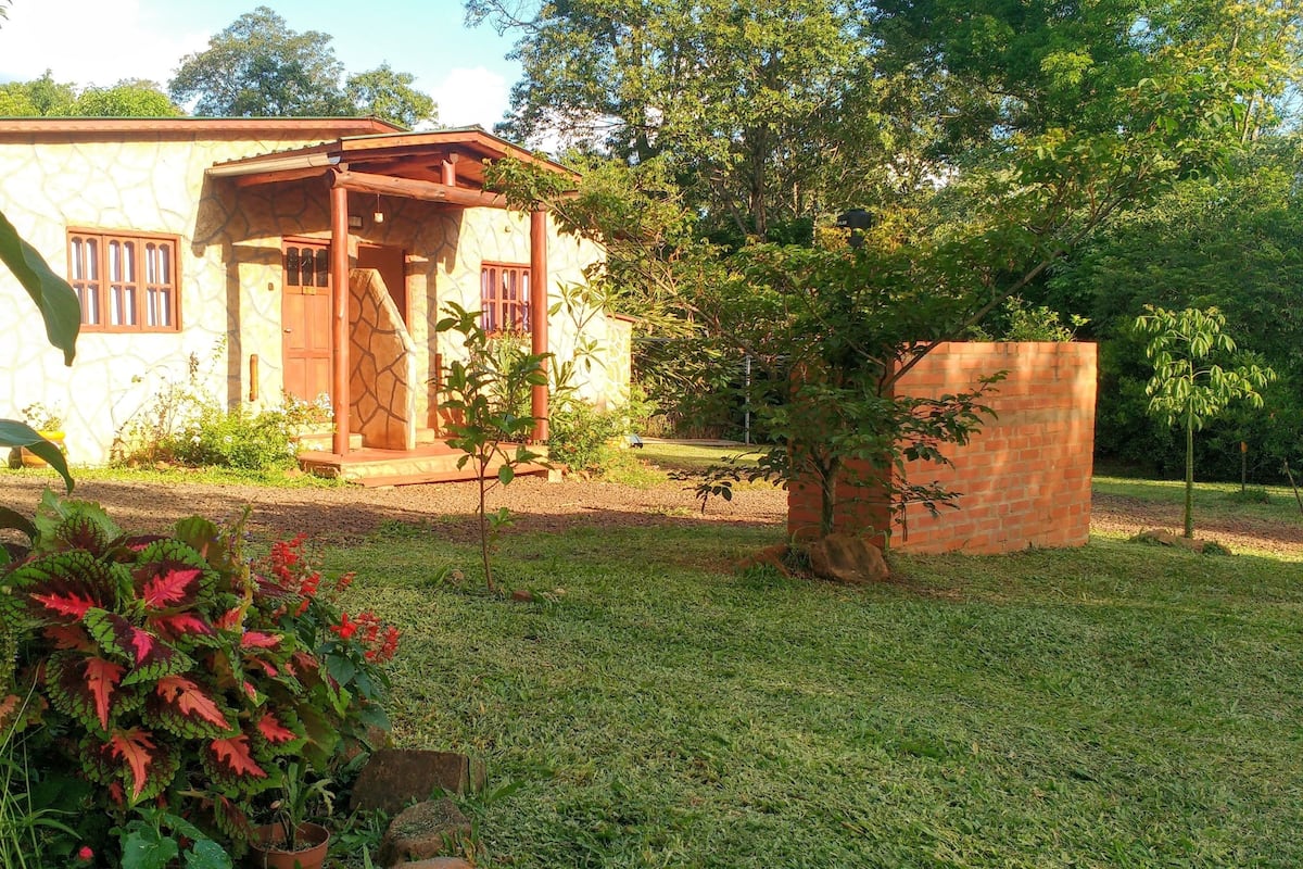 A house with a stone facade, a wooden porch, and a well-maintained garden with various plants and a small tree.