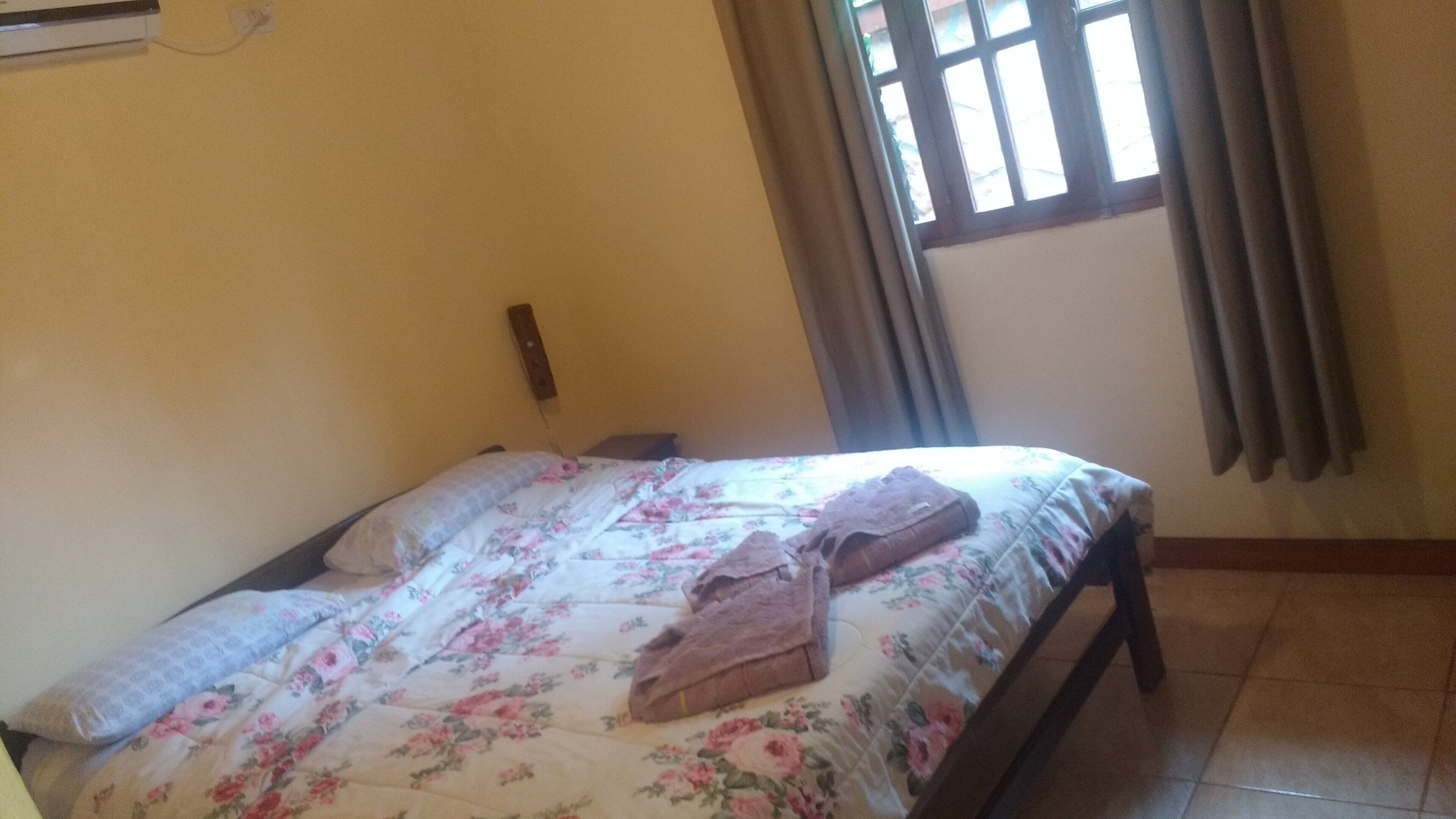 Ein Schlafzimmer mit einem Bett, floralem Bettbezug, einem Fenster mit Vorhängen und einer Wandklimaanlage.
