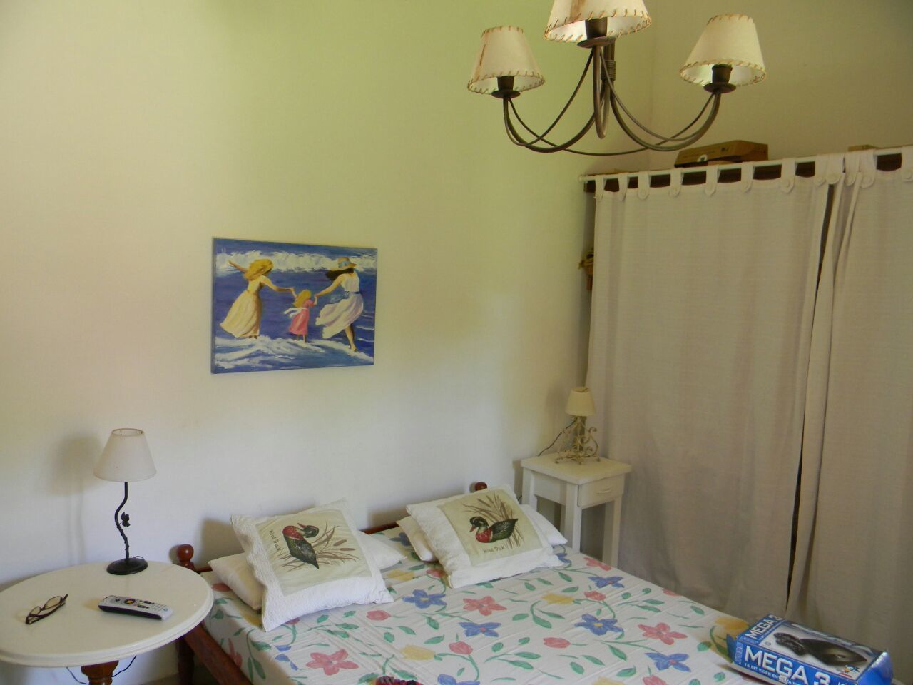 A bedroom with a bed, a nightstand, a table with a lamp, a painting on the wall, and a window with curtains.
