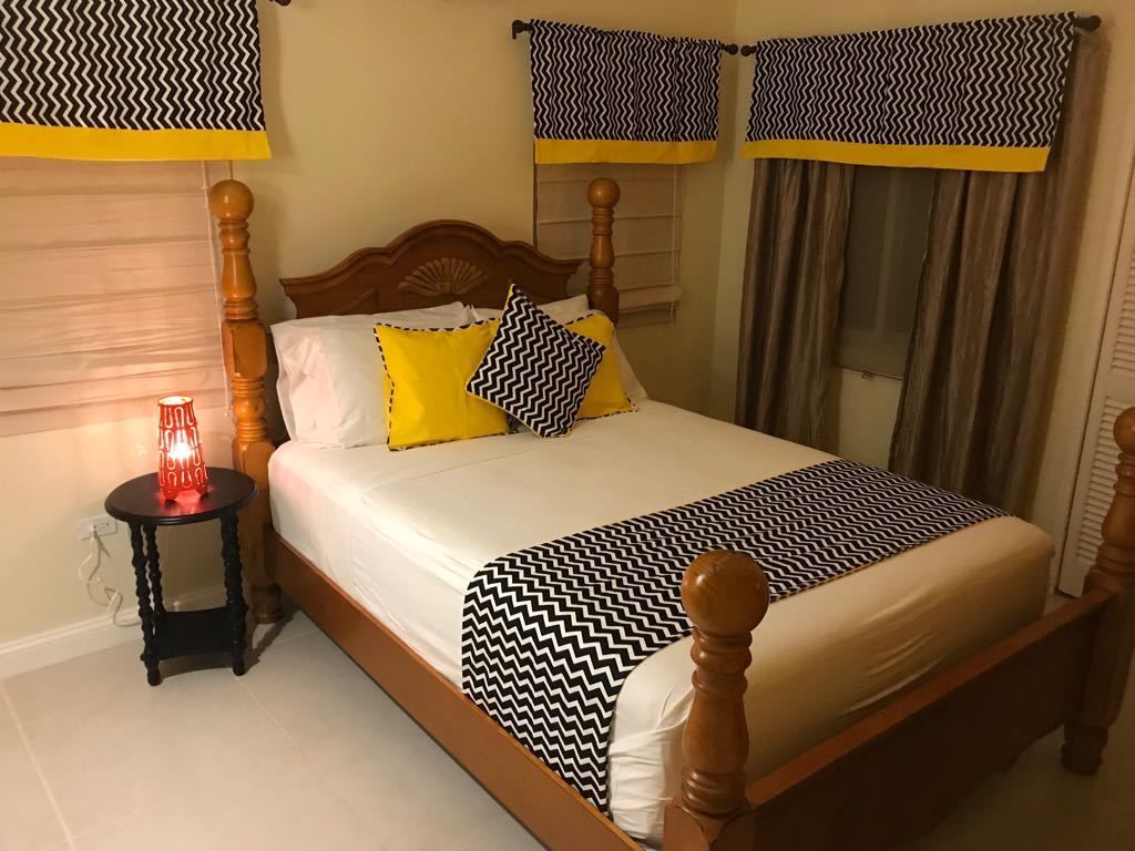 Un lit bien fait, recouvert d’une couvre-lit à motifs, une table de chevet avec une lampe rouge et des rideaux à rayures en zigzag.