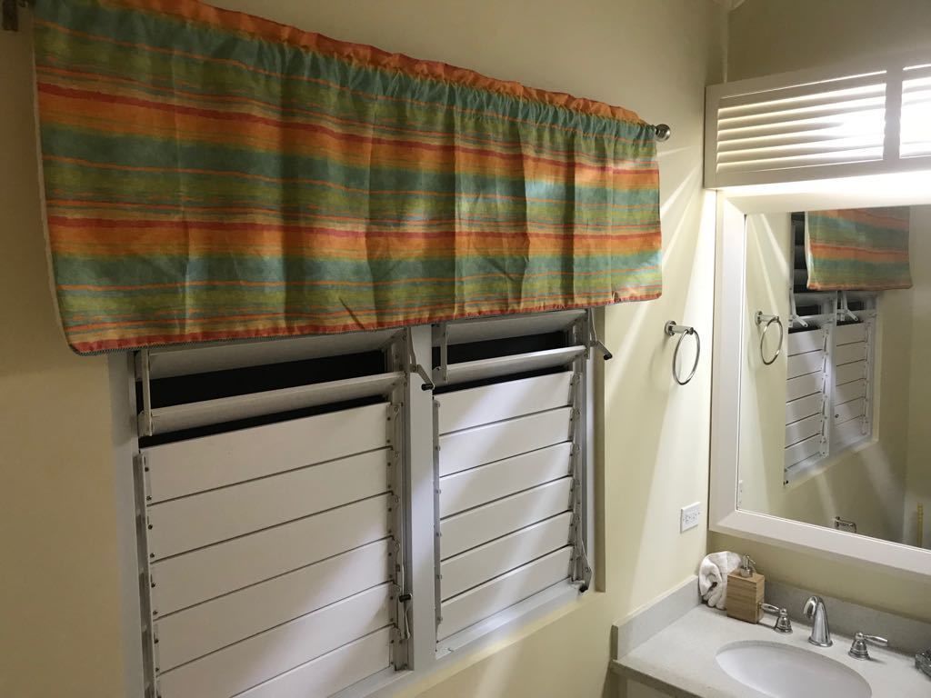 Un baño con ventana cubierta por una cortina a rayas, un lavabo con espejo y un perchero para toallas.