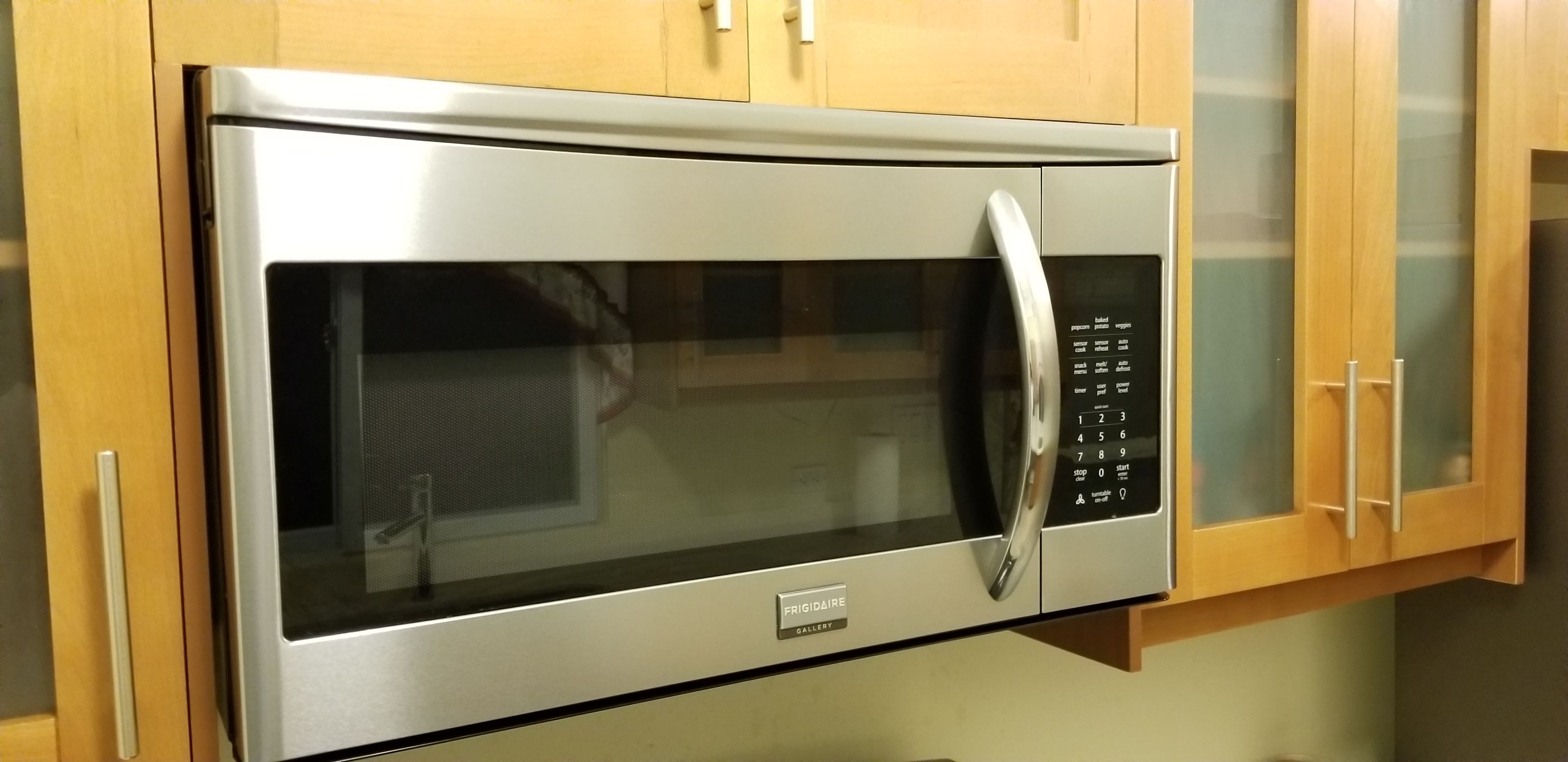 Un four à micro-ondes en acier inoxydable doté d’un panneau de commande numérique, fixé sur un meuble en bois avec des portes en verre.