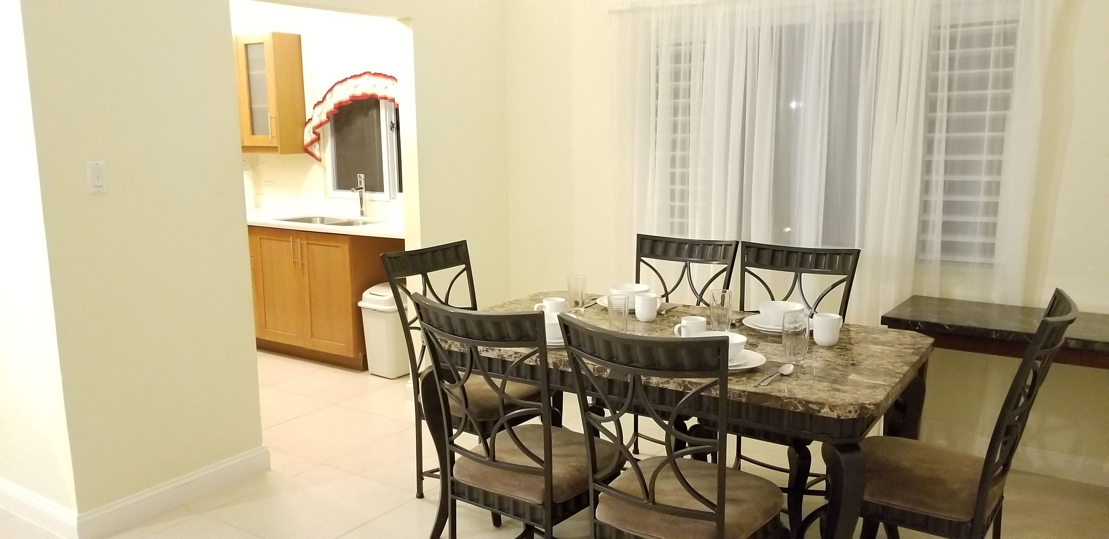A dining area with a marble table and four chairs, a kitchen in the background, and a window with sheer curtains.