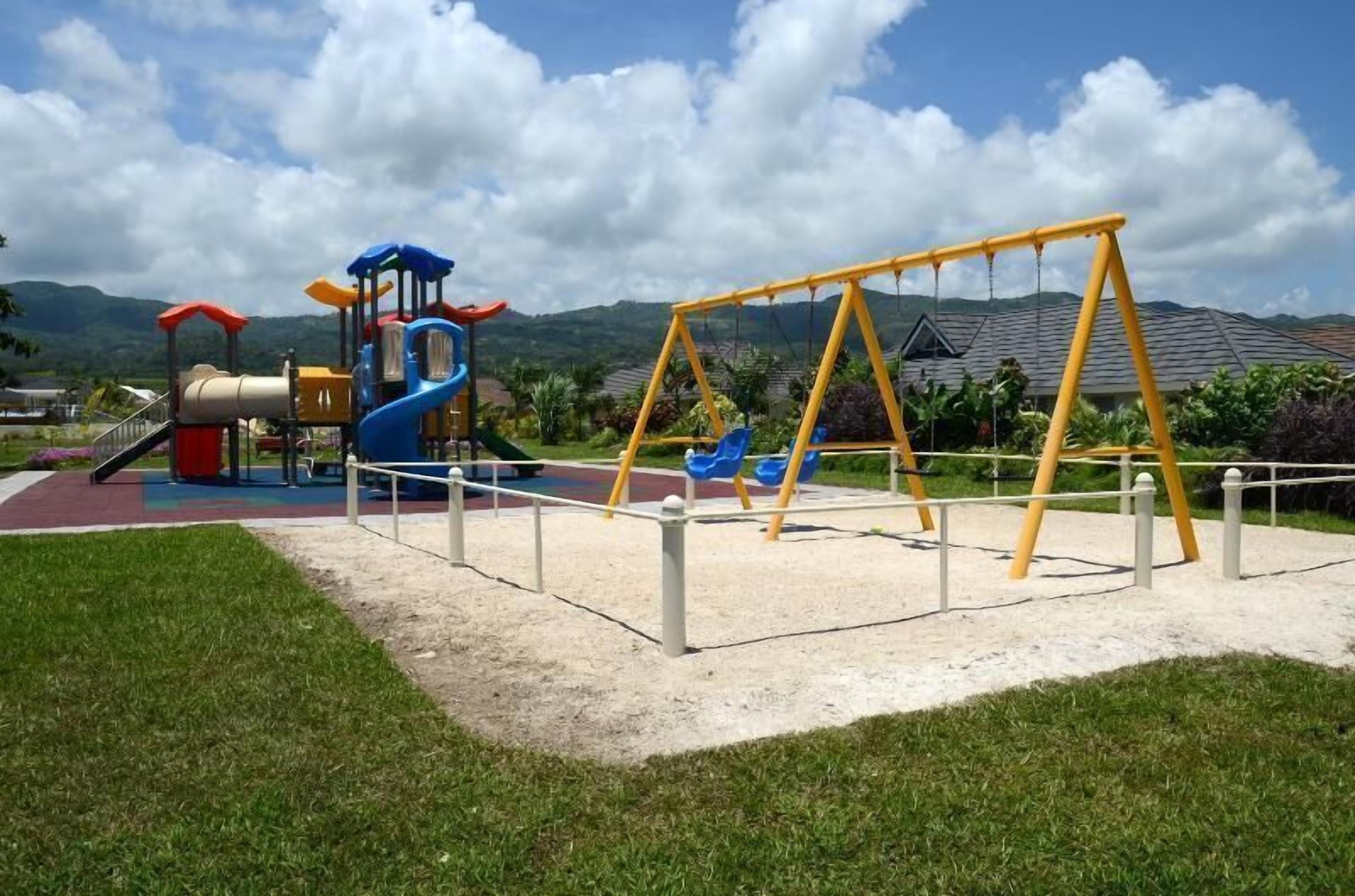 Un terrain de jeux avec des balançoires et des toboggans, entouré de pelouse et bordé d’un paysage vallonné.