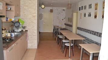 A kitchen with a dining area, featuring a long table and chairs, a stove, and various kitchen utensils.