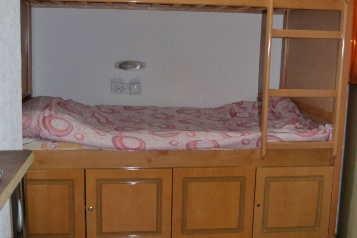 Una cama individual doble con escritorio y silla debajo, un taburete pequeño y suelo de baldosas.