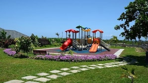 Un parque infantil con toboganes y una estructura para escalar, rodeado de un jardín con flores moradas y un sendero de piedra.