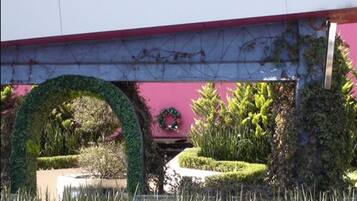 A building with a pink facade, a covered walkway, and a well-maintained garden with a circular hedge and various plants.