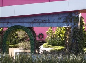 Un edificio con fachada rosa, un paseo techado y un jardín bien cuidado con setos circulares y diversas plantas.