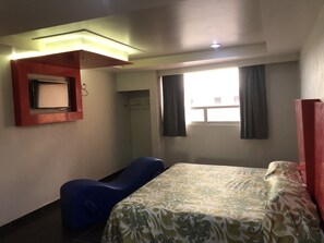 Habitación de hotel con cama, una silla azul, una mesita, ventana con cortinas y televisor montado en la pared.
