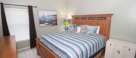 A bedroom with a bed, a wooden dresser, a nightstand, a lamp, and a window with curtains.