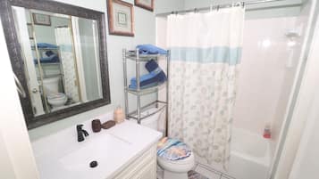 Un baño con un lavamanos blanco, un espejo, un inodoro con una cubierta de asiento colorida, una cortina de ducha y un perchero.