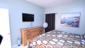 Une chambre à coucher avec un lit, une commode, un téléviseur, un ventilateur et un tableau accroché au mur.
