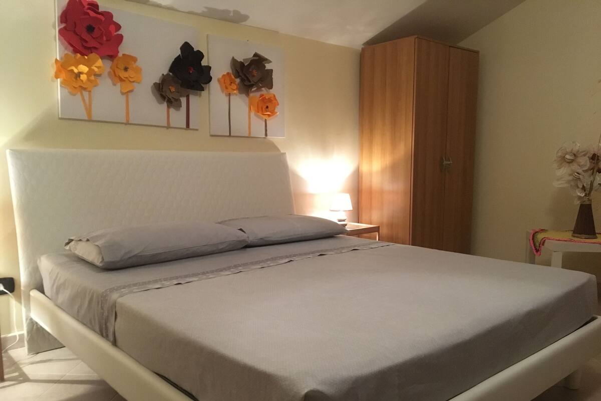 A neatly made bed with a grey bedspread and pillows, a wooden wardrobe, and a wall with three flower paintings.