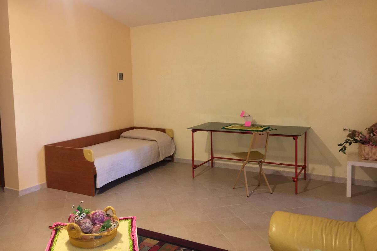 A small, cozy room with a single bed, a desk with a chair, and a small table with a basket of fruit.