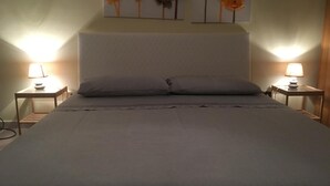 Una cama bien hecha con un colchón y almohadas grises, flanqueada por dos mesitas de noche con lámparas, y una obra de arte en la pared.