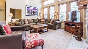 Una sala de estar acogedora con un sofá modular, una mesa de centro y un televisor. Amplias ventanas proporcionan luz natural y vistas a árboles.