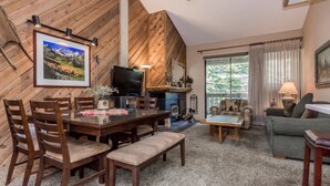 A cozy living room with a wooden wall, a fireplace, a television, and a dining area with chairs and a bench. - Crestview B46 (Mammoth Lakes)