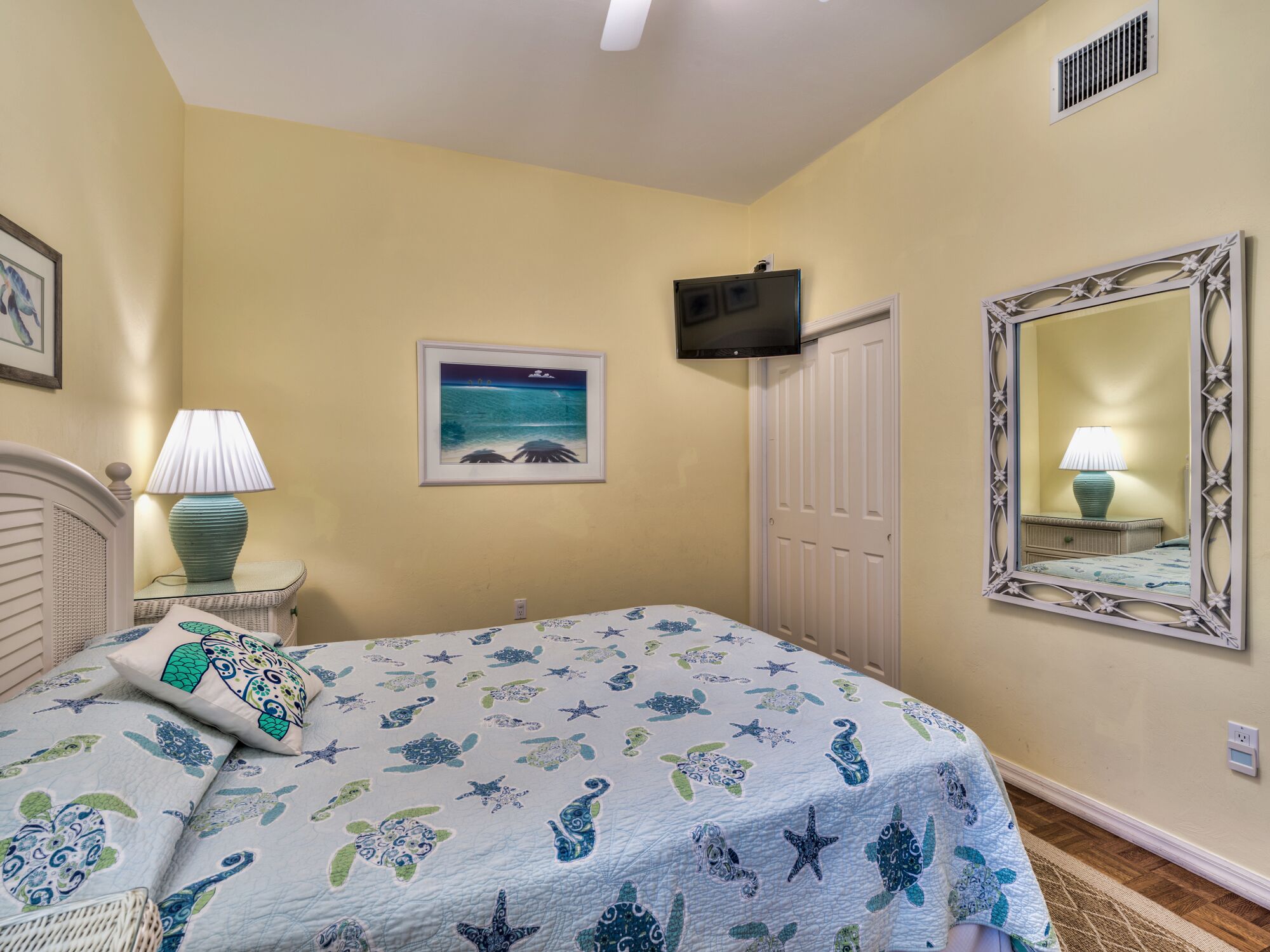 A bedroom with a bed, a nightstand, a lamp, a television, a door, a framed picture, and a mirror.