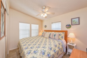A bedroom with a bed, a ceiling fan, two windows, a nightstand, and a wall-mounted lamp.