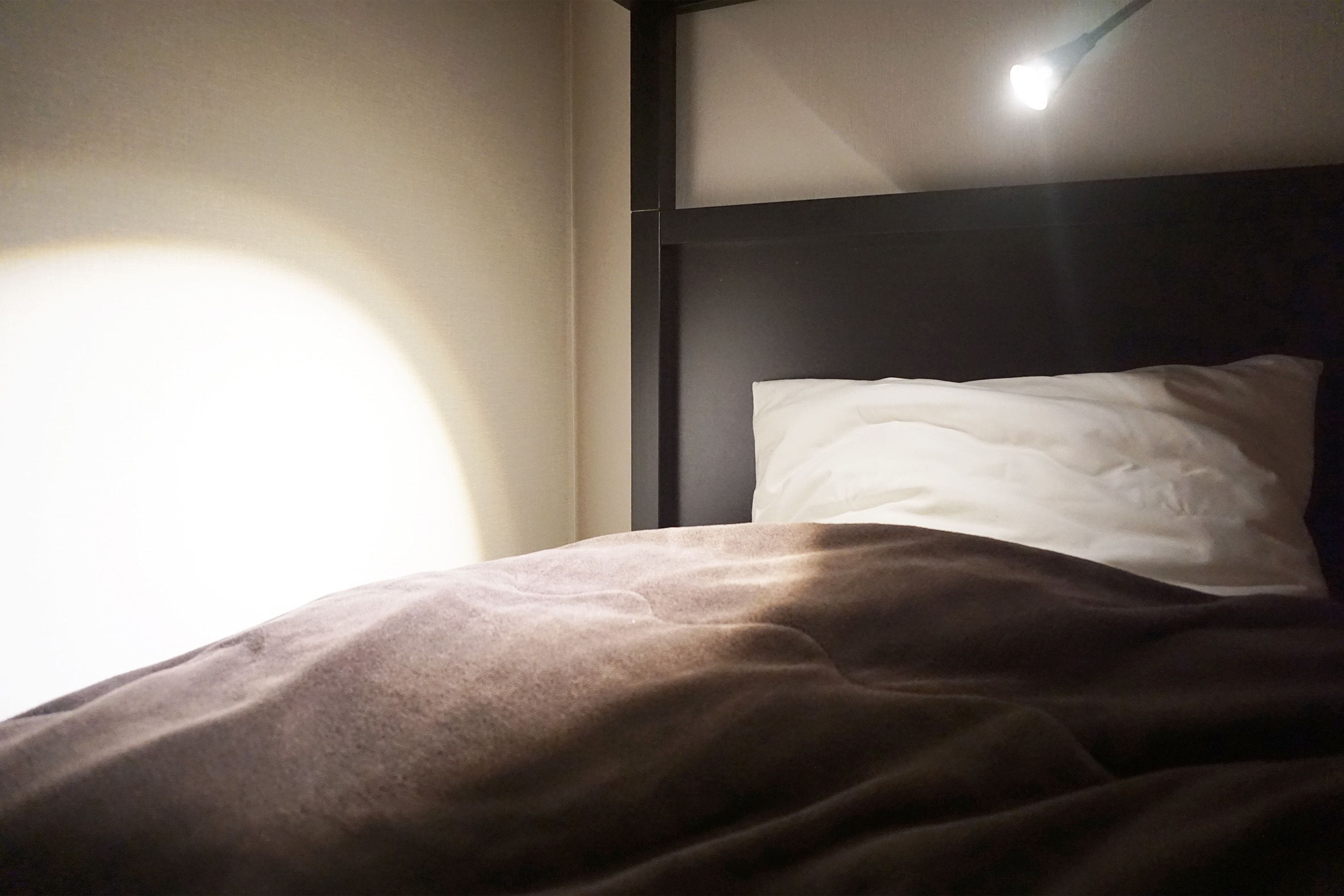 Ein Bett mit einem weißen Kopfkissen und einer braunen Decke in einem Zimmer, das ein dunkles Kopfteil und eine Lichtquelle darüber hat.