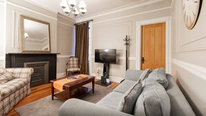 A living room with a sofa, armchair, coffee table, television, fireplace, and a clock on the wall. - Elegant 3 Bed Apt ★ ★ ★ ★ ★ Mins to Manhattan (Hoboken)