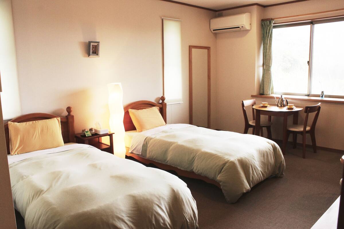 A hotel room with two beds, a desk, a chair, a window with curtains, and an air conditioning unit.