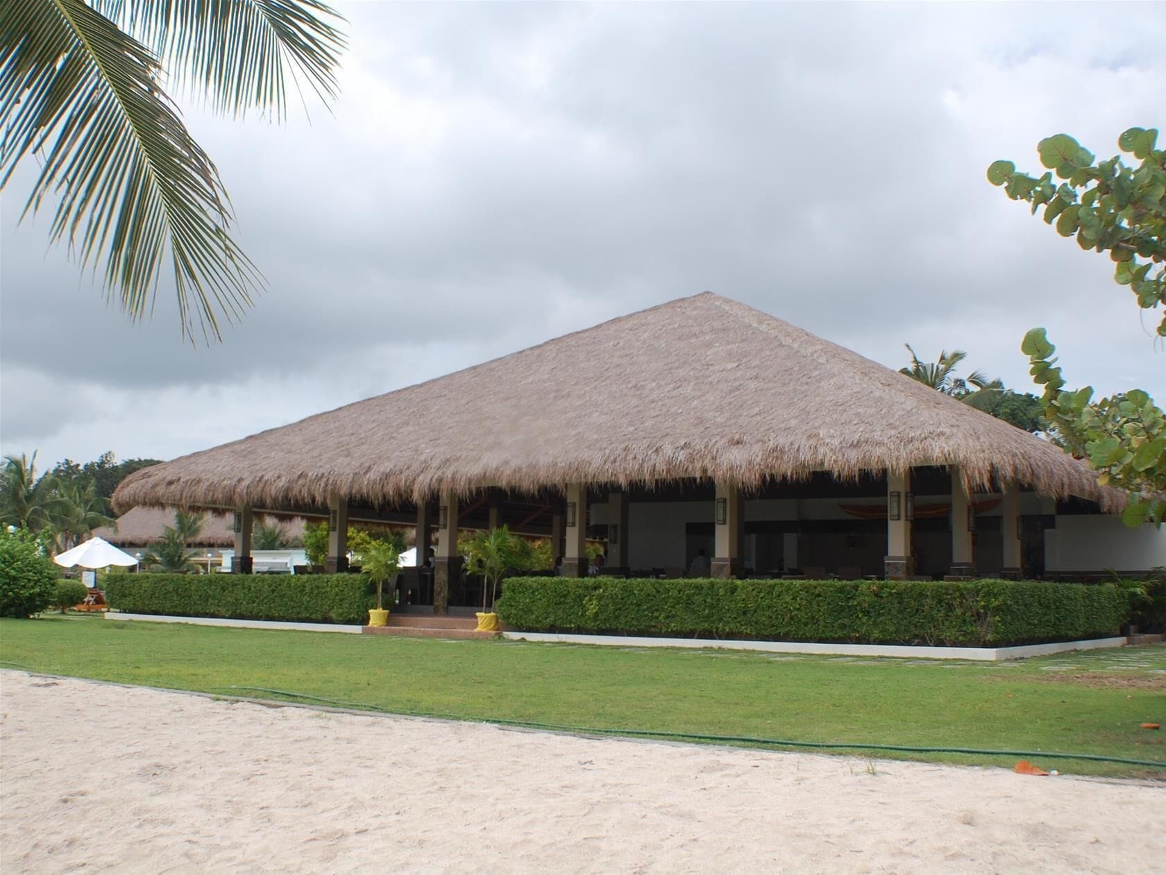 A thatched-roof building with a covered outdoor area, surrounded by well-maintained greenery and a sandy foreground.