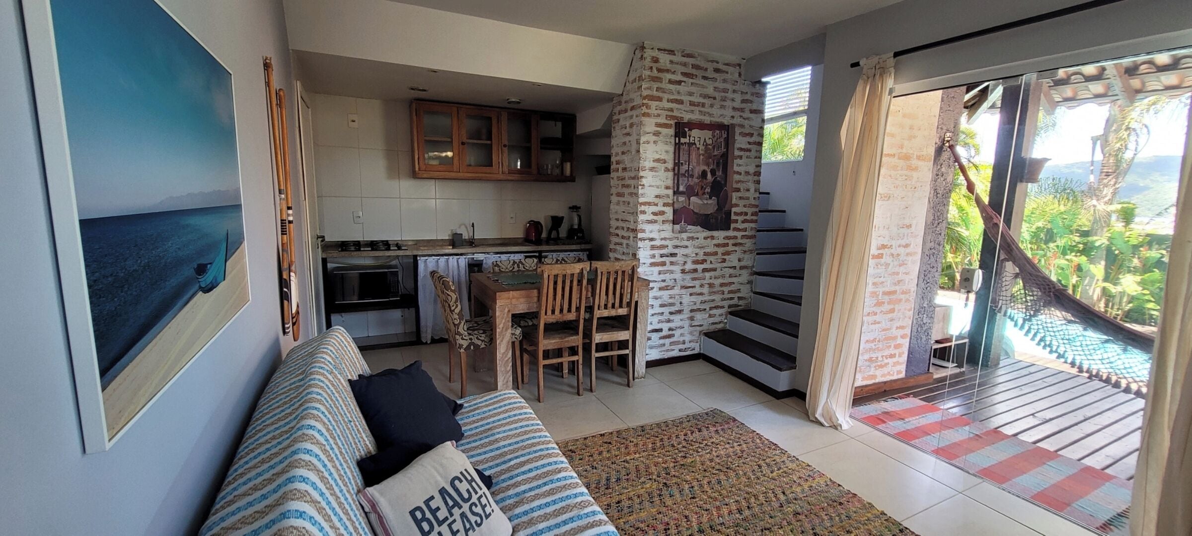 Een gezellige woonkamer met een bank, een eethoek met stoelen, een keuken met kasten, en een trap die leidt naar een balkon met uitzicht.