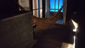 Un pasillo tenuemente iluminado con una hamaca y una escalera que lleva a una habitación con una ventana iluminada en azul.