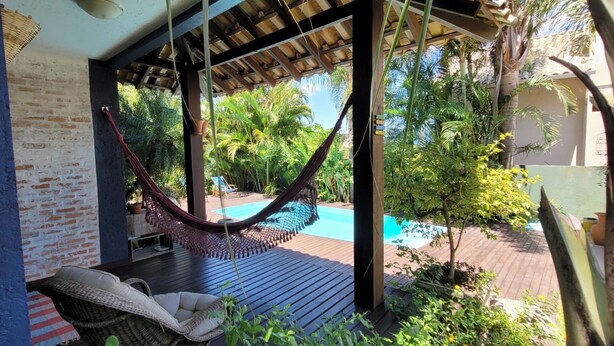 Una hamaca bajo una pérgola de madera, una piscina y abundante vegetación.